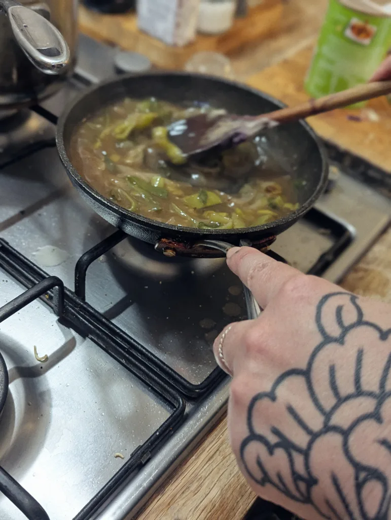 Sliced leeks in a brown sauce simmering in a frying pan on a gas hob, stirred with a wooden spoon by a person with a large hand tattoo.