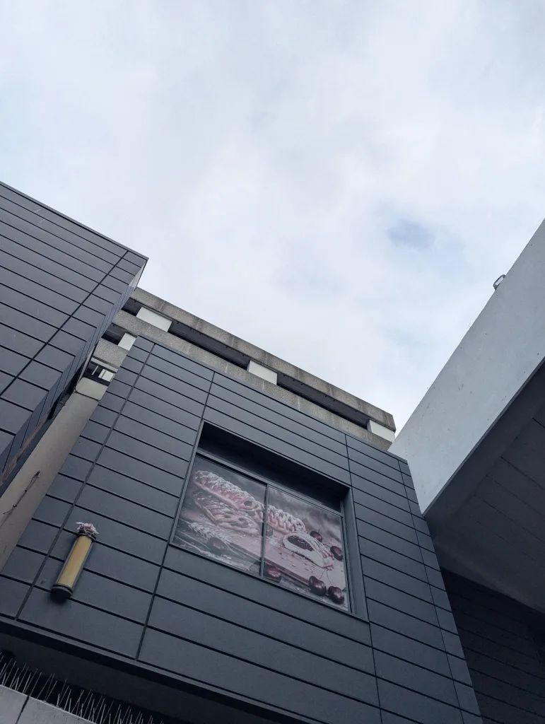 Dark grey panelled wall with a window showing cherry pastries, seen from a low angle against concrete architecture and a cloudy sky.