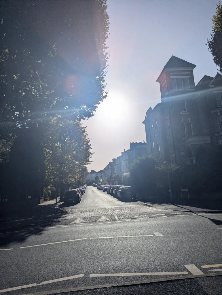 Sunlight flares over a residential street lined with tall trees and parked cars, featuring a large gabled brick house on the right.