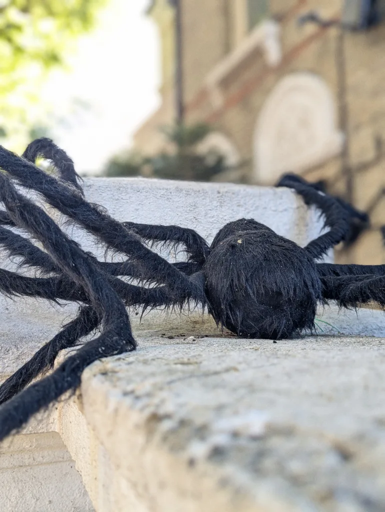 A large, fuzzy black fake spider decoration resting on a stone ledge with a blurred brick building in the background.