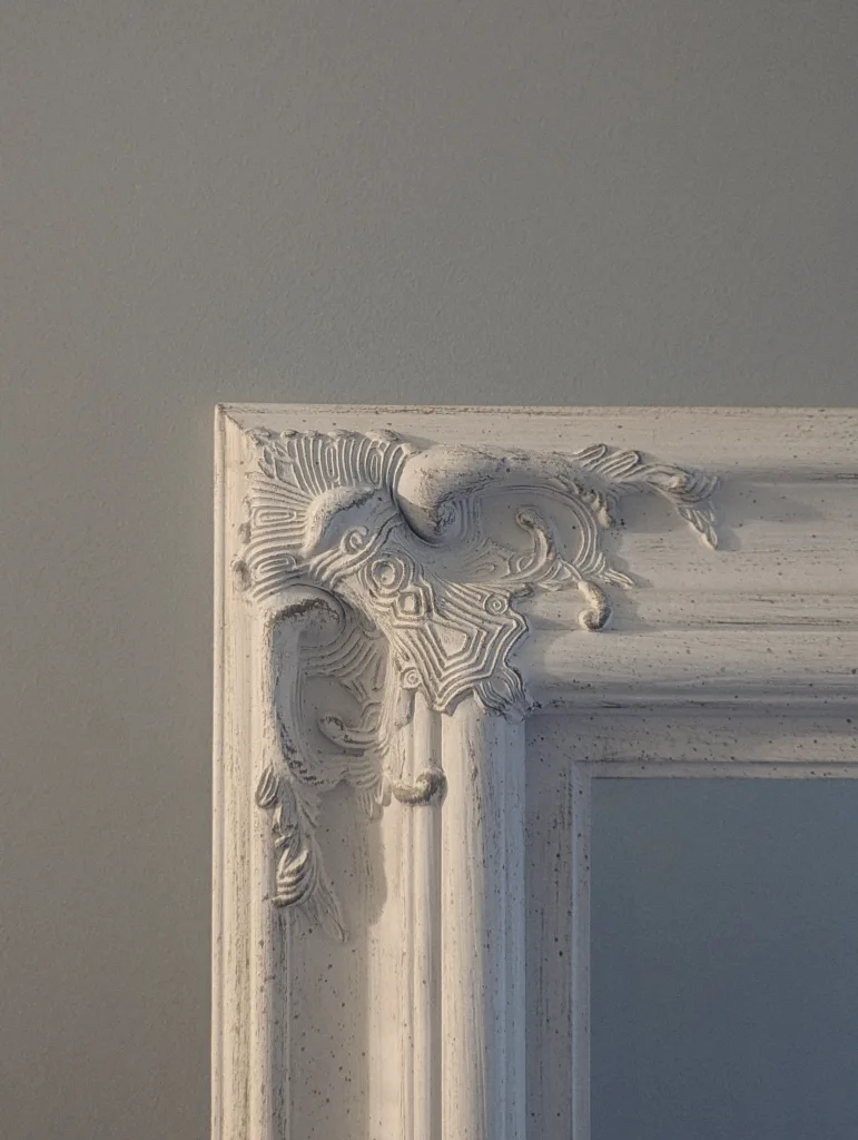 The upper left corner of an ornate white mirror frame with intricate scrollwork and a weathered finish against a plain grey wall.
