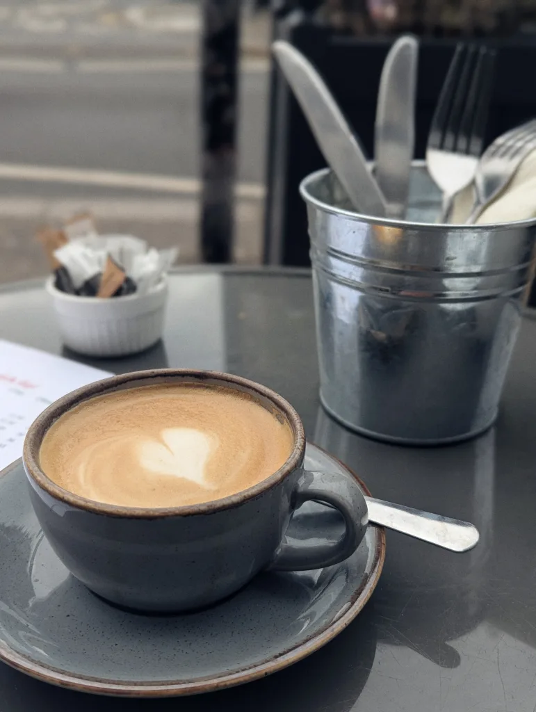 Heart-shaped latte art in a grey ceramic cup on an outdoor café table beside a metal bucket of cutlery and sugar sachets.