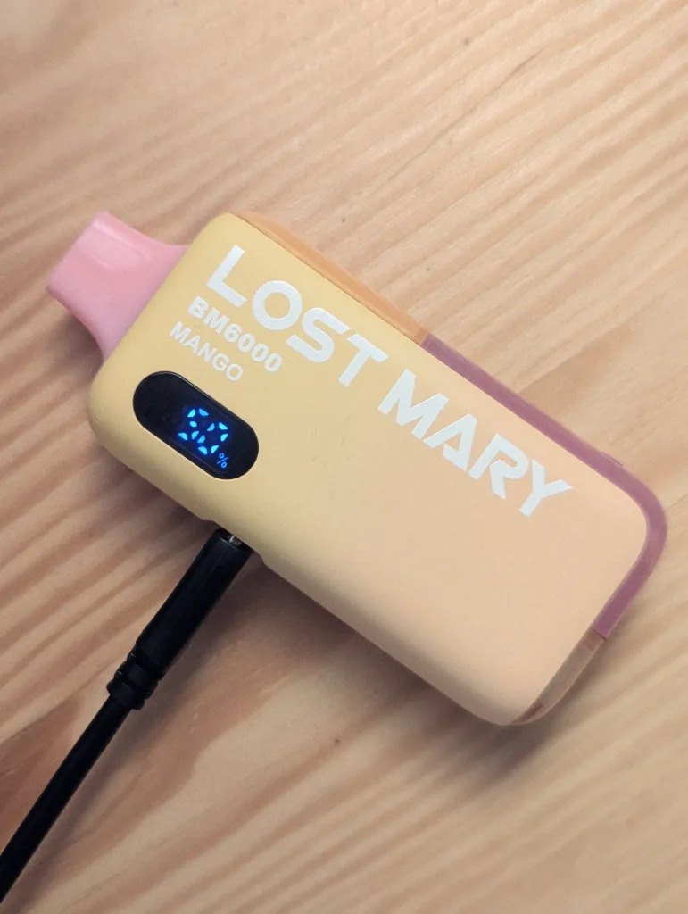 A yellow Lost Mary BM6000 Mango vape charging on a wooden surface, its digital screen displaying 50% battery.