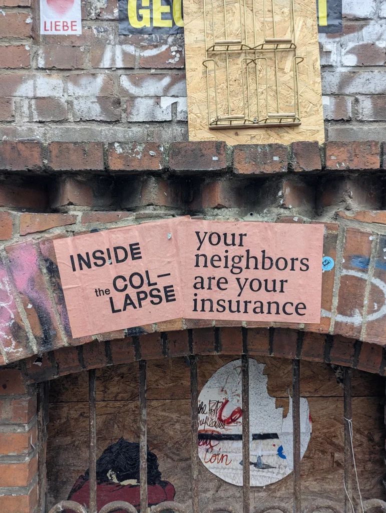 Pink posters on a brick archway reading 'INSIDE the COL-LAPSE your neighbors are your insurance' above a basement window with rusty bars.