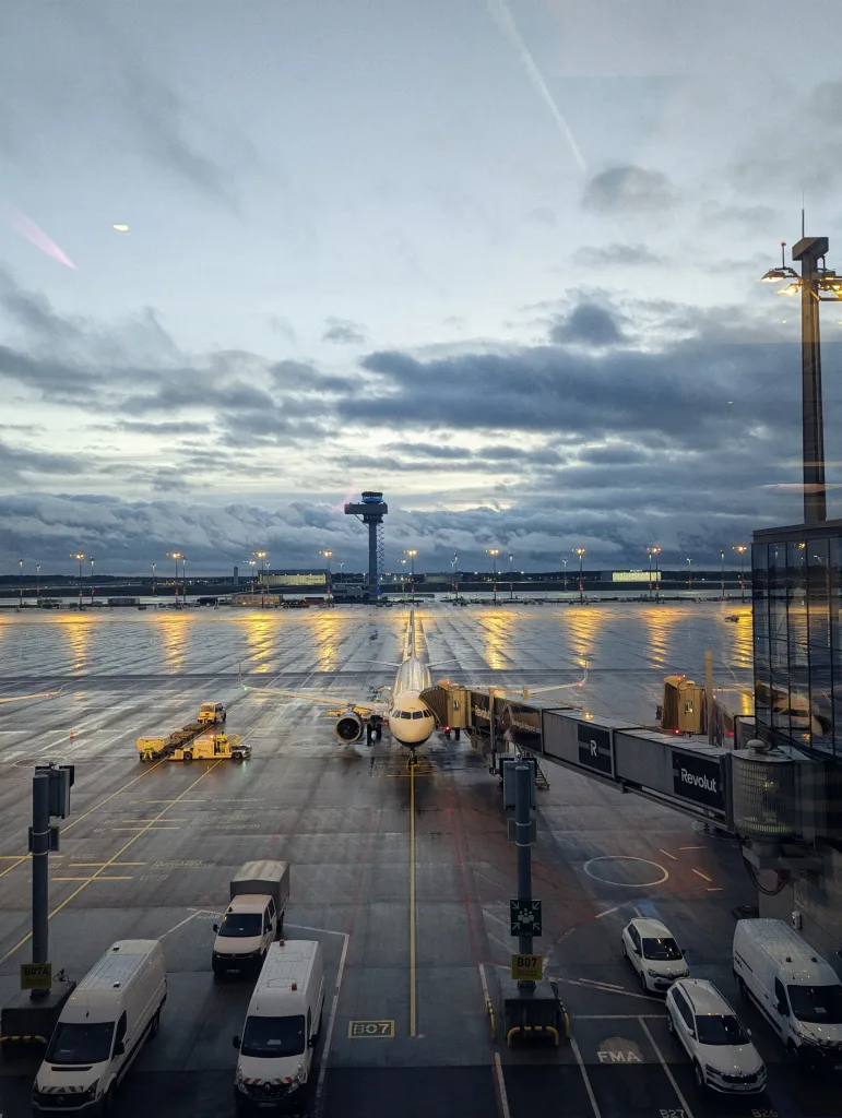 An airplane at a terminal gate on wet tarmac at dusk. Reflections of lights on the ground under dramatic clouds and a distant control tower.