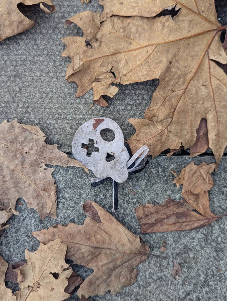 A small white cardboard skull with a cross-shaped eye lying on stone pavement among dry, brown autumn leaves.