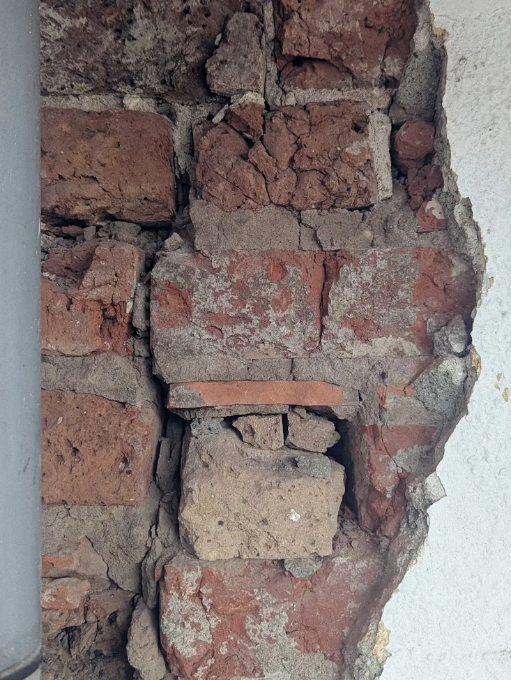 Exposed, crumbling red bricks and mortar revealed behind a jagged hole in a white plastered wall next to a grey pipe.