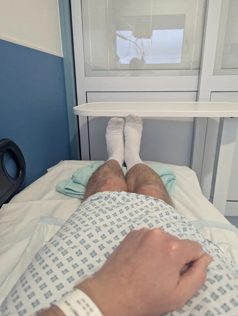 First-person view from a hospital bed showing legs in white socks, a blue-patterned gown, and a patient ID wristband on the person's wrist.