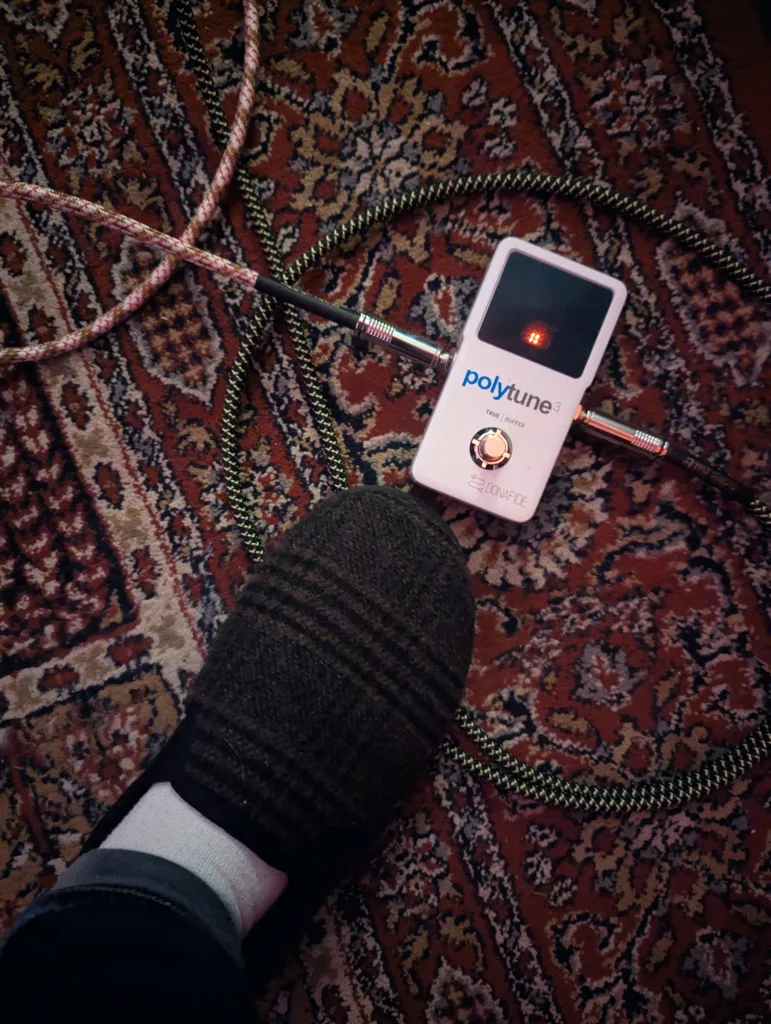 A white PolyTune 3 guitar tuner pedal with braided cables on a red patterned rug, next to a foot in a brown plaid slipper.