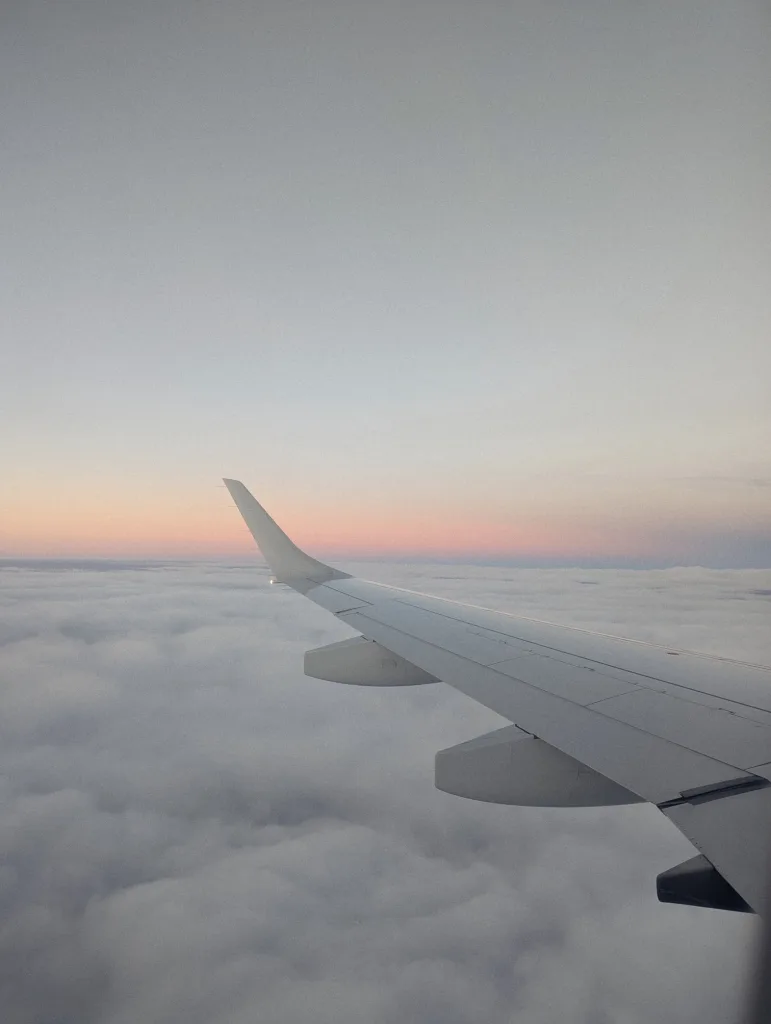 An airplane wing extends over a thick layer of white clouds beneath a soft pink and orange sunset horizon under a pale sky.