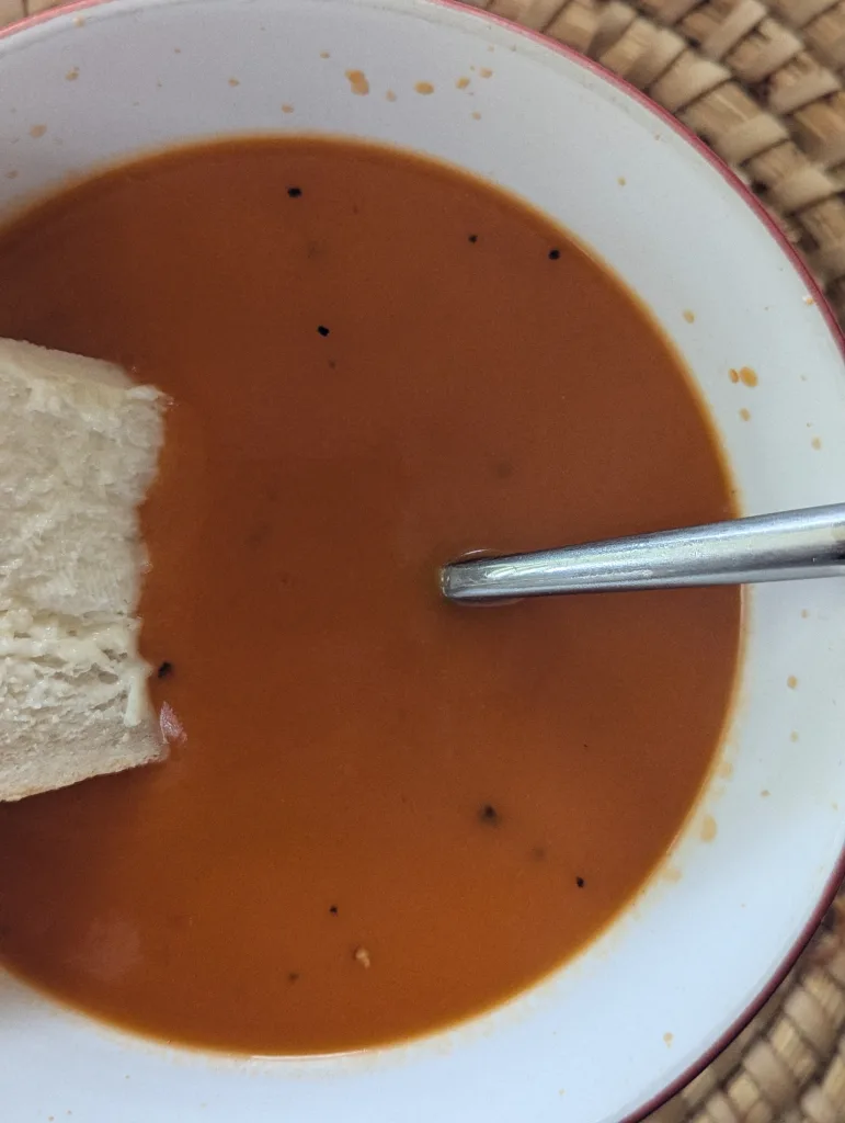 Smooth tomato soup with black pepper in a red-rimmed white bowl, served with crusty bread and a silver spoon on a woven mat.