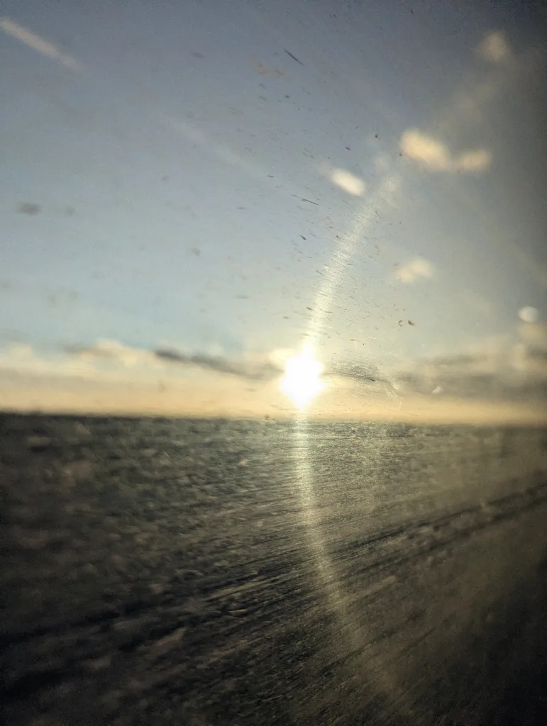 Bright sun on the horizon viewed through a scratched glass window, creating a vertical lens flare against a pale blue sky.