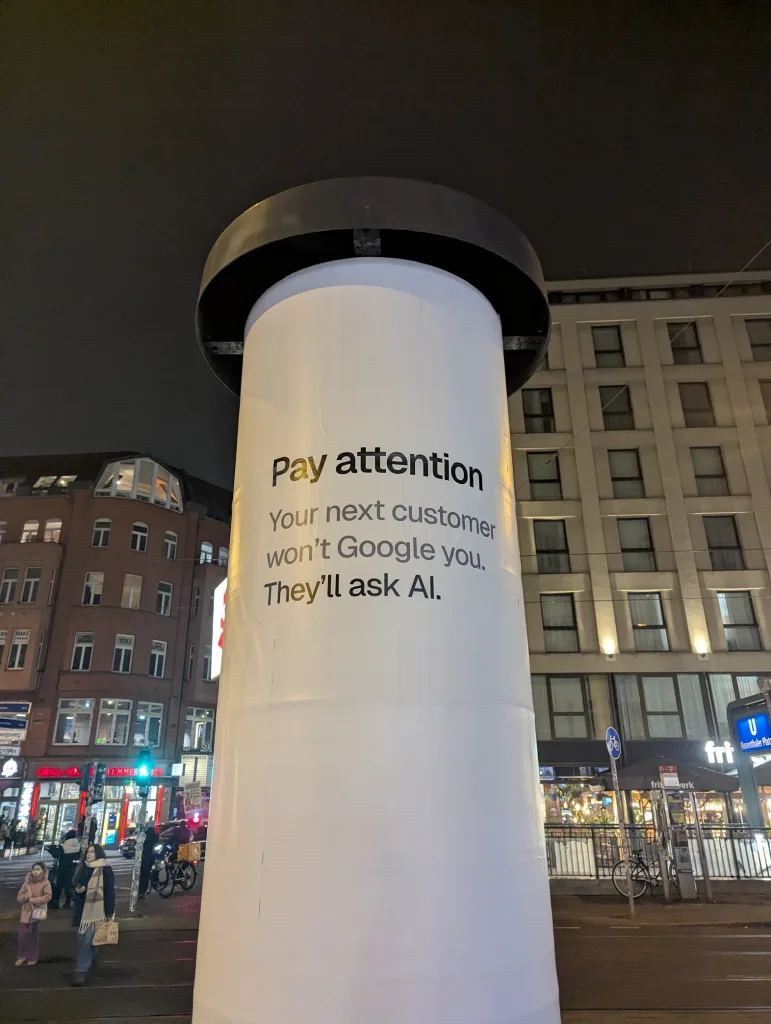 A white advertising pillar at night in a city street reading: Pay attention Your next customer won't Google you. They'll ask AI.