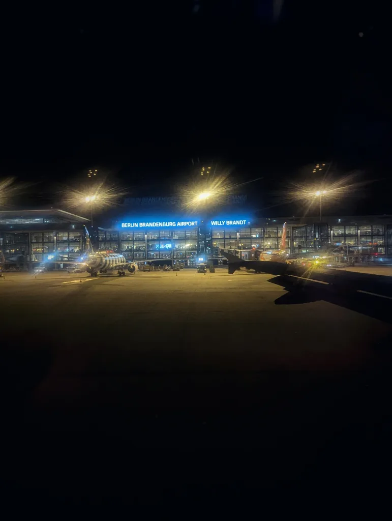 Berlin Brandenburg Airport at night with glowing blue signage, aircraft on the tarmac, and bright floodlights casting lens flares.