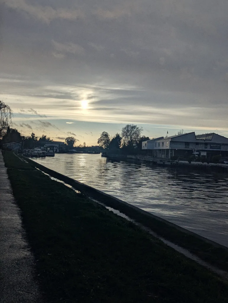 Hazy sun setting over a river, reflecting on the water between a grassy bank and white riverside buildings under a cloudy sky.