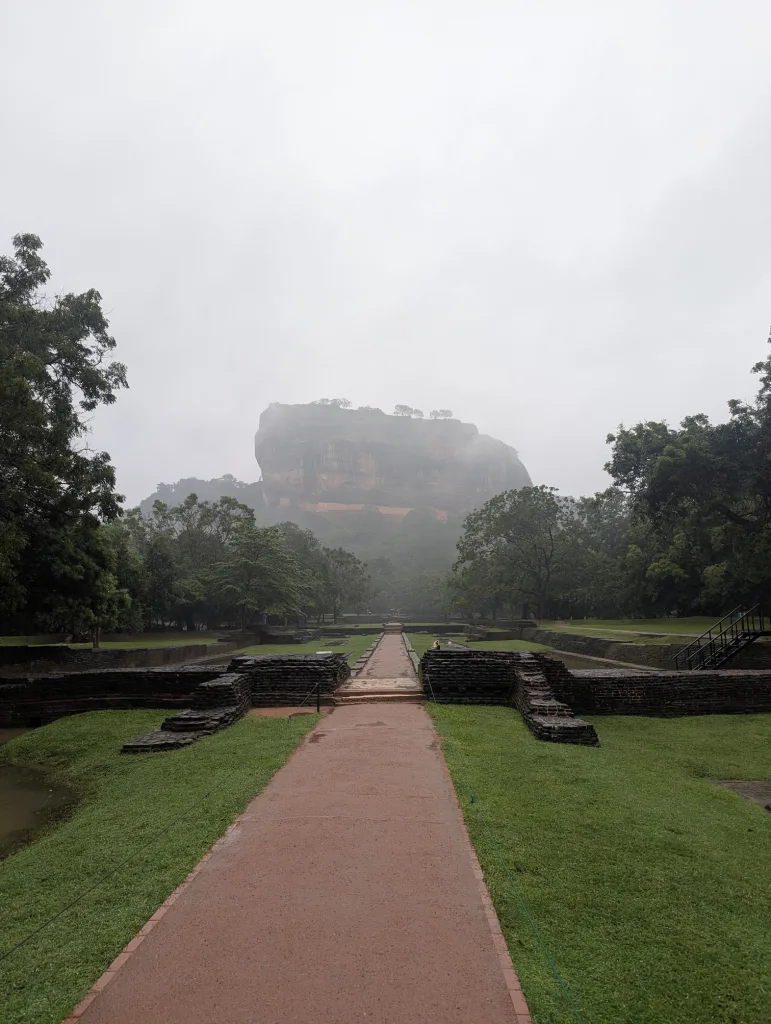 A straight red pathway leading towards the misty Sigiriya rock fortress, flanked by green lawns and ancient stone foundations.