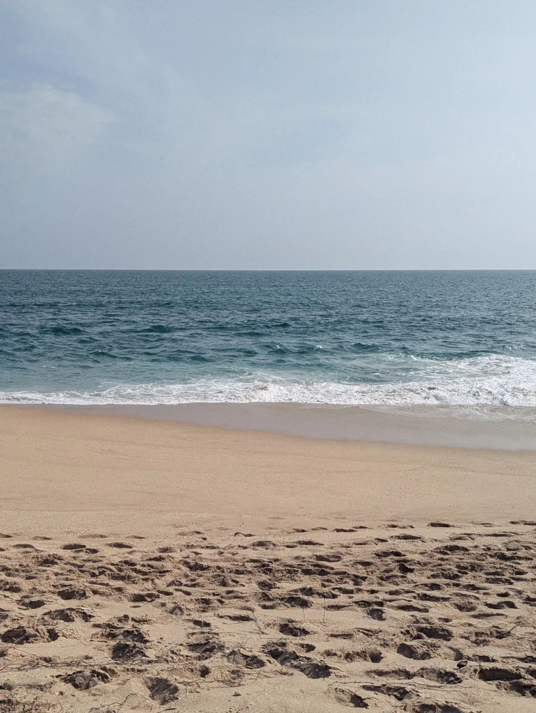 Footprints in golden sand leading to white waves on a deep blue ocean under a clear, pale blue sky.