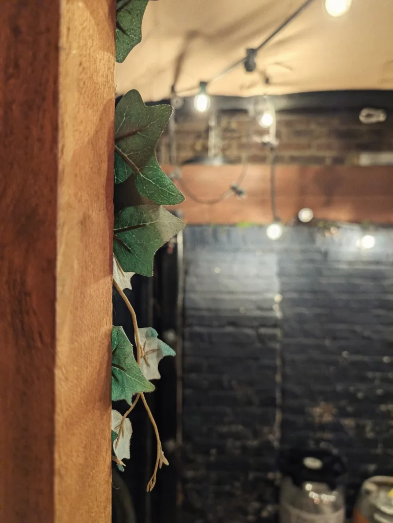 Artificial ivy leaves on a wooden pillar against a blurred background of glowing string lights and a dark painted brick wall.