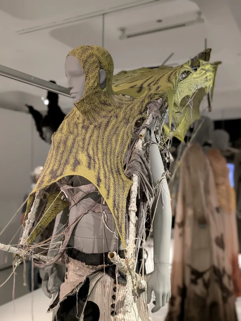 Gray mannequin in a gallery wearing a yellow and black knit hooded garment with wing-like extensions held by white branch-like frames.