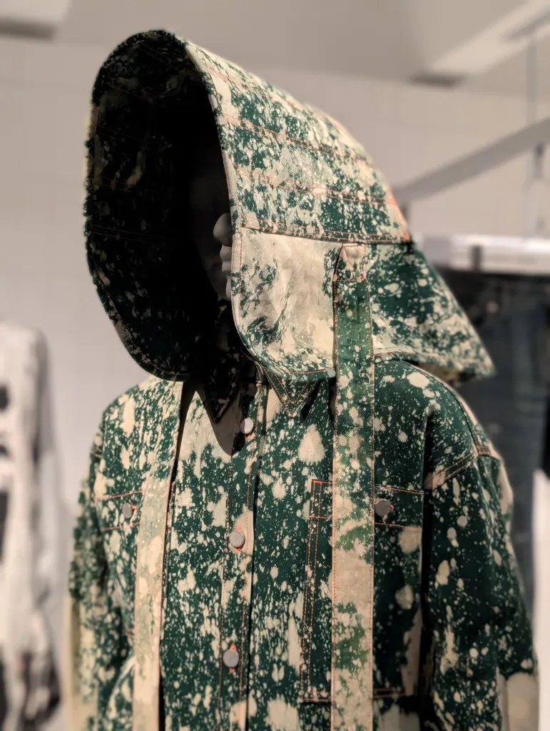 A mannequin wearing a green denim jacket with cream bleach splatters and a distinctive, wide structured hood in a retail display.