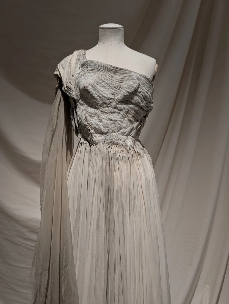 Beige gown on a mannequin featuring a textured, encrusted bodice, a one-shoulder drape, and a finely pleated skirt against draped fabric.