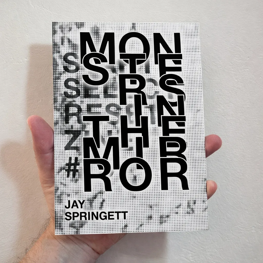 Monsters in the Mirror Zine by Jay Springett - a hand holding a book with bold black typography on a white halftone background.