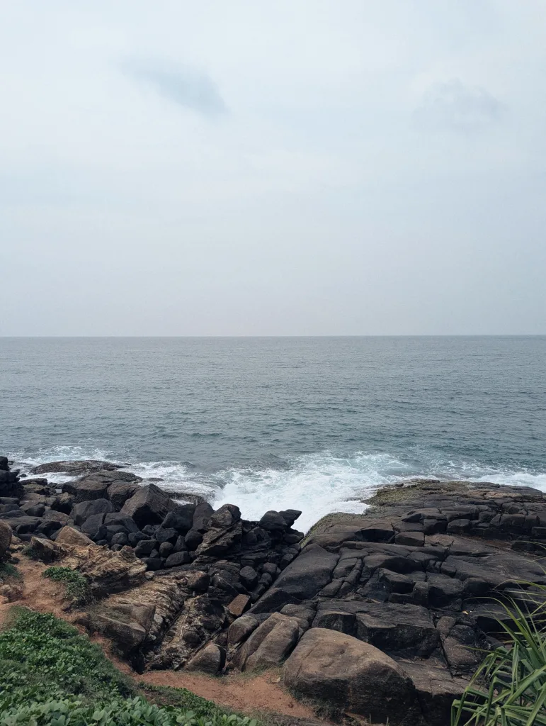Dark, jagged rocks along a coastline as ocean waves create white foam under an expansive, hazy sky.