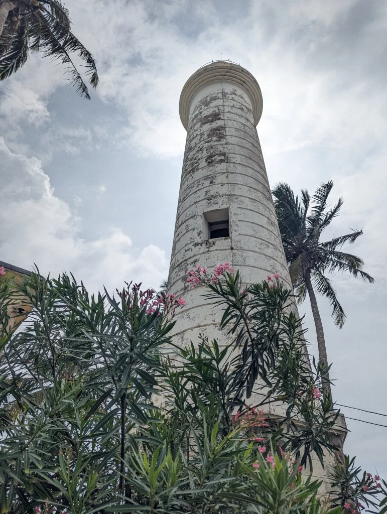 A tall, weathered white lighthouse towers behind flowering pink oleander bushes and palm trees against a cloudy sky.