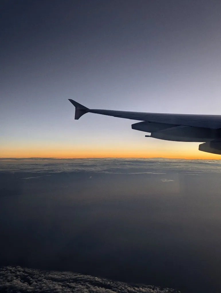 An airplane wing silhouetted against a sunset horizon over a layer of clouds, with a gradient sky fading from orange to deep blue.