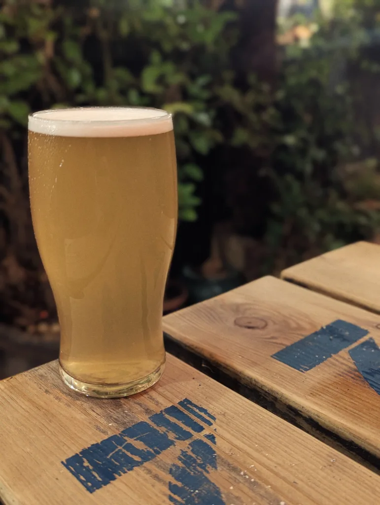 A hazy golden pint of beer with a frothy head sits on a wooden table with blue stencilled markings, in front of lush green foliage.