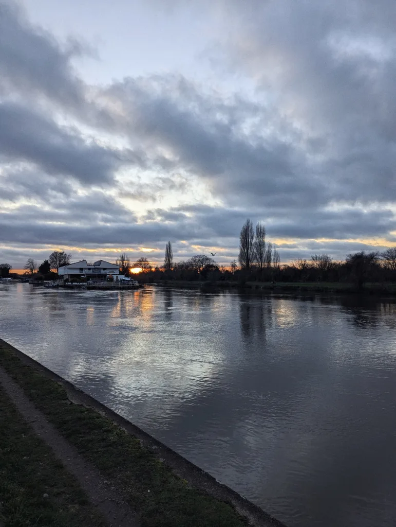Sunset glow breaking through dark clouds over a wide river, with a white building and tall trees on the bank.