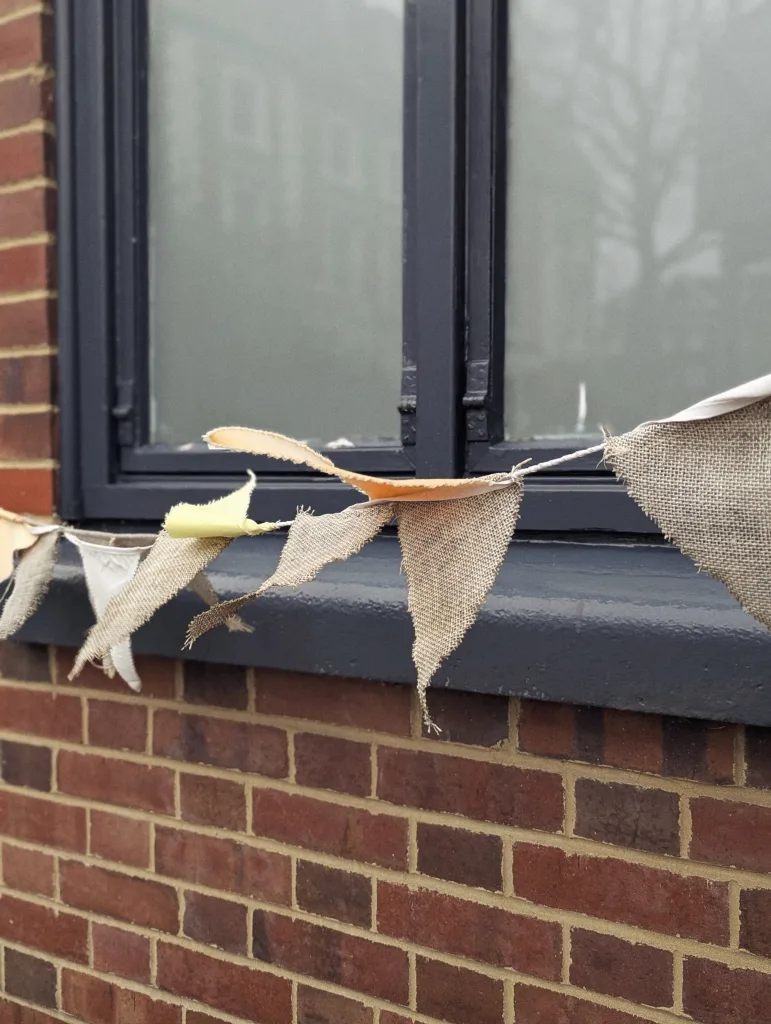 Frayed burlap and cloth bunting hanging across a dark grey window frame against a red brick wall.