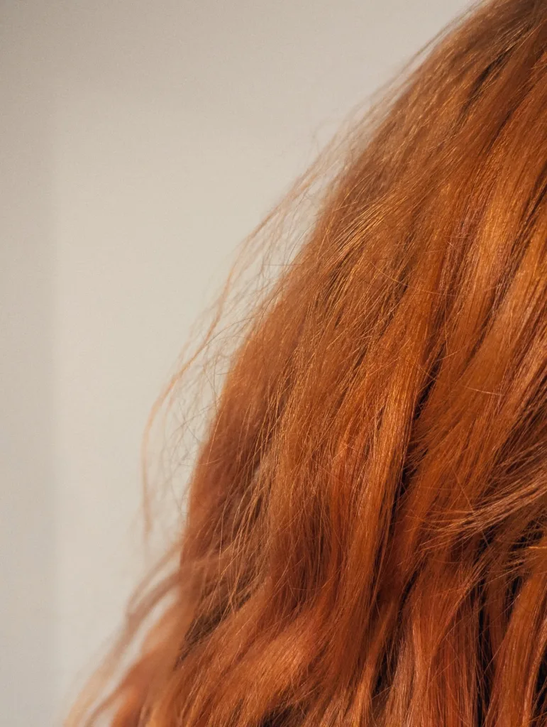 Close-up of vibrant orange-red wavy hair with flyaway strands against a neutral off-white background.