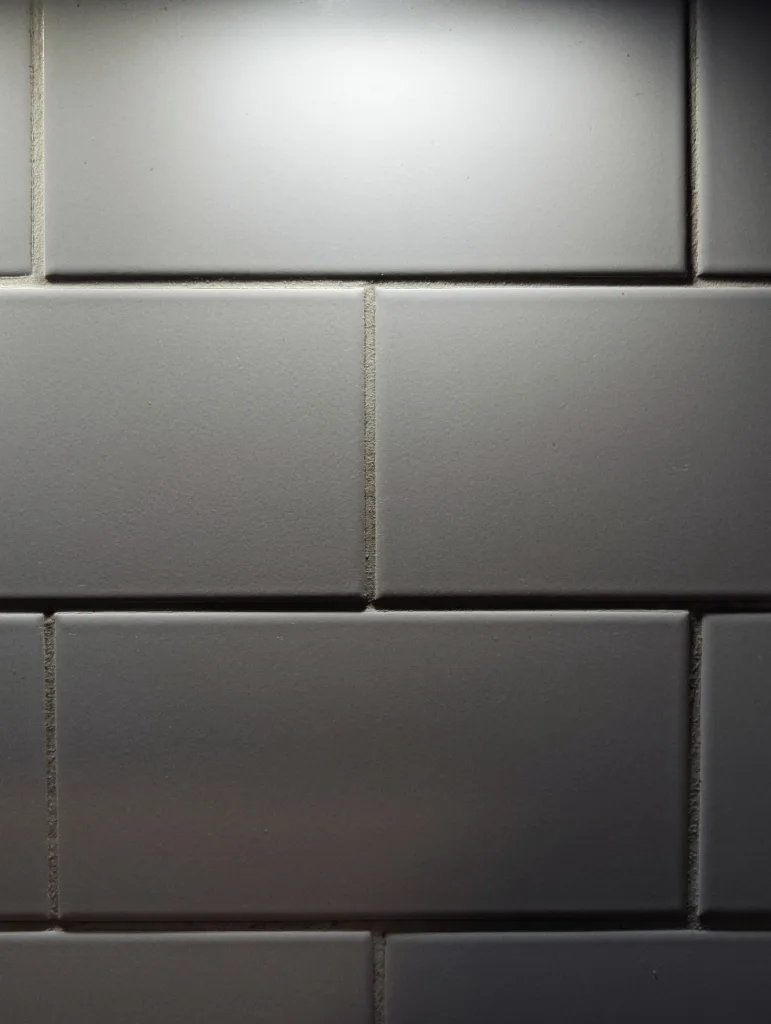 Grey rectangular wall tiles in a brick pattern, with overhead light casting shadows into the recessed grout lines.