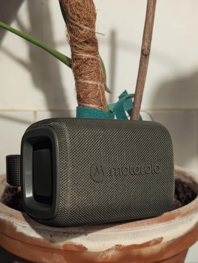A grey mesh Motorola speaker rests in a terracotta plant pot next to a climbing plant's moss pole and a wooden support stake.