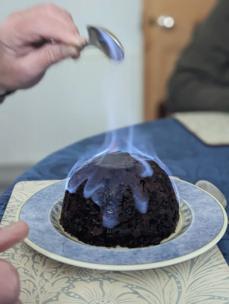 A dark Christmas pudding flambéed with bright blue flames on a blue plate, with a hand holding a flaming spoon over the top.
