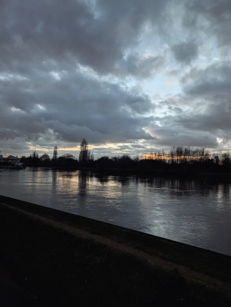 Dramatic sunset over a wide river with heavy grey clouds and orange light on the horizon, silhouetted trees lining the far bank.