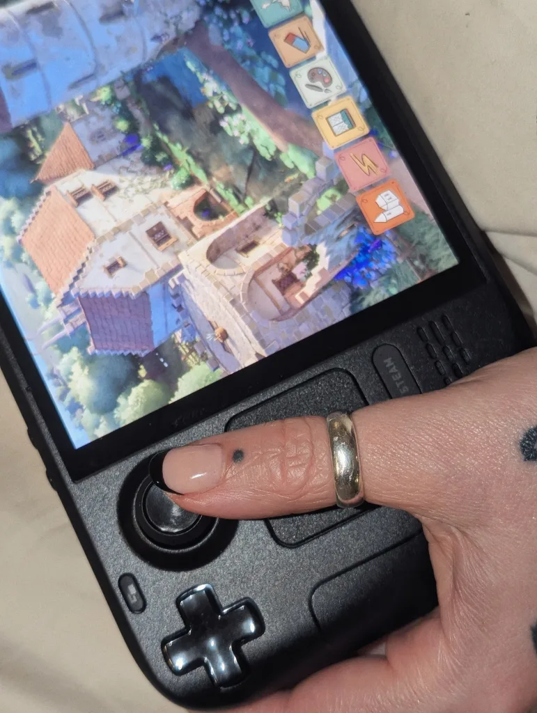A hand with a silver ring and black tipped nails playing Tiny Glade on a Steam Deck handheld console.