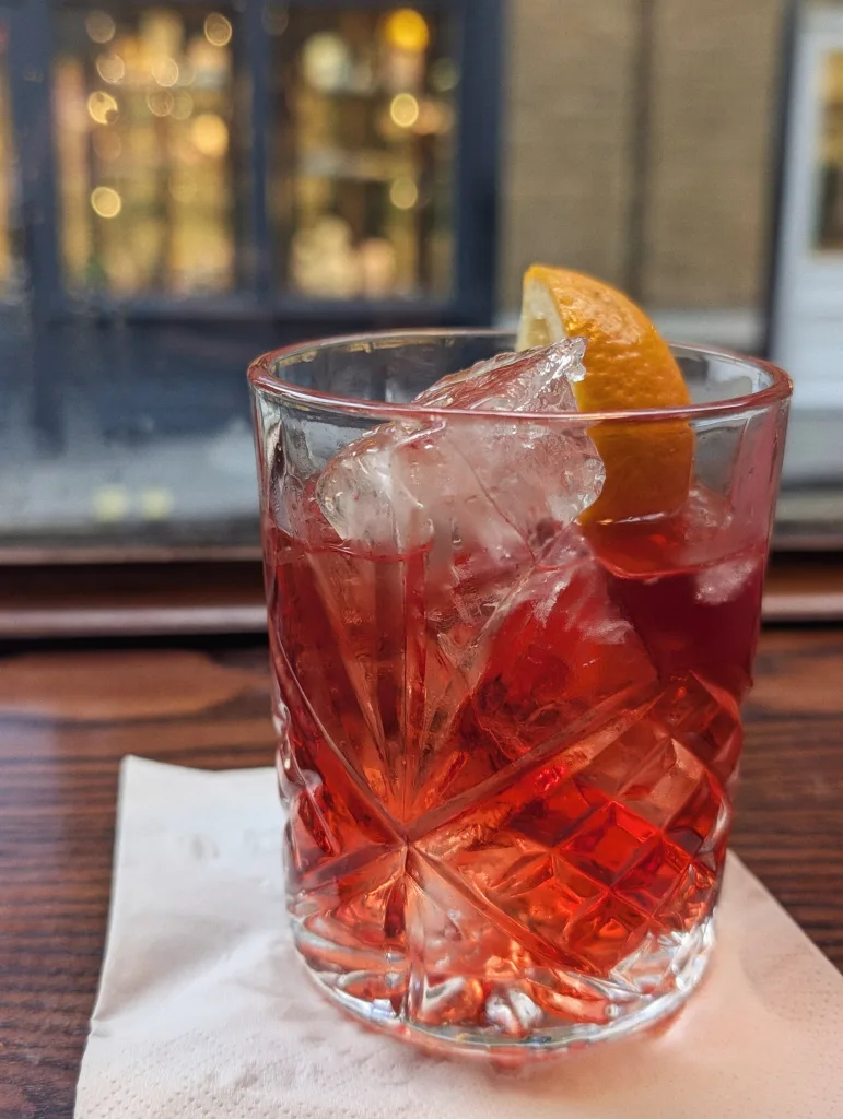 Red negroni cocktail with ice and an orange slice in a cut crystal glass on a white napkin, against a background of blurred golden lights.