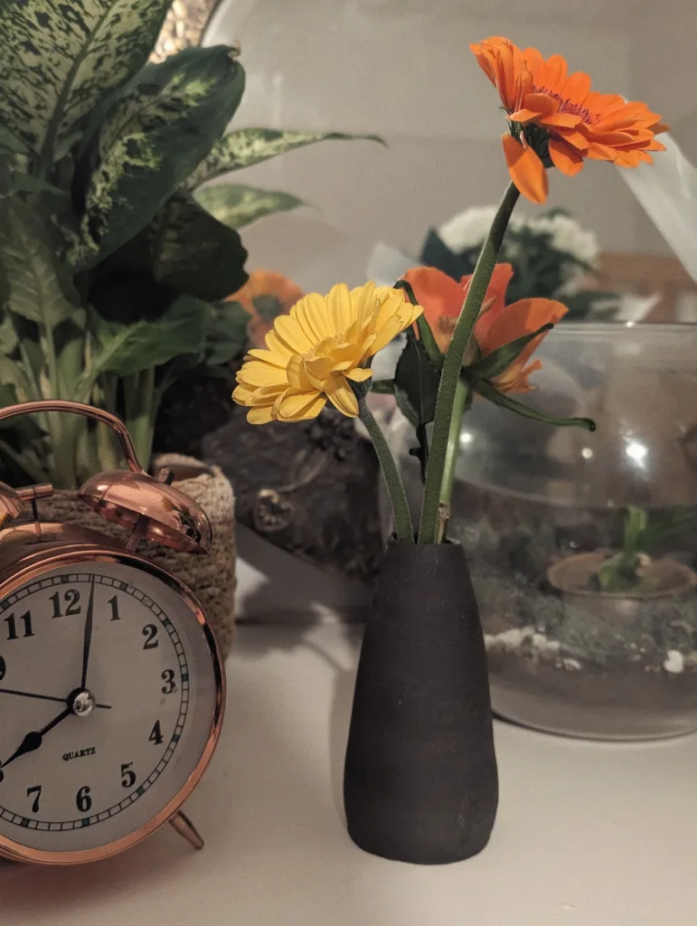 A copper alarm clock beside a small black vase holding yellow and orange gerbera daisies, with green leafy plants in the background.