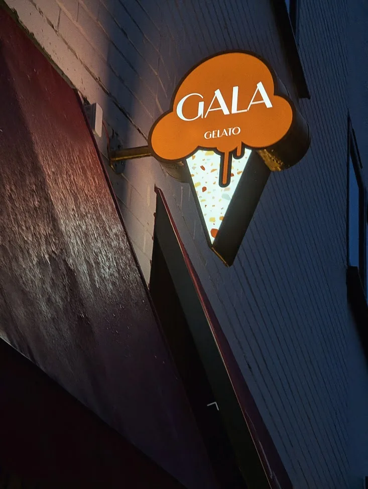 An illuminated Gala Gelato sign shaped like an ice cream cone with an orange scoop and a speckled cone mounted on a white brick wall at night.