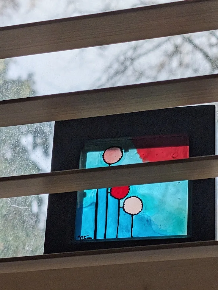 Stained glass art with stylized pink and red flowers on a teal background, viewed through horizontal wooden blinds against a window.