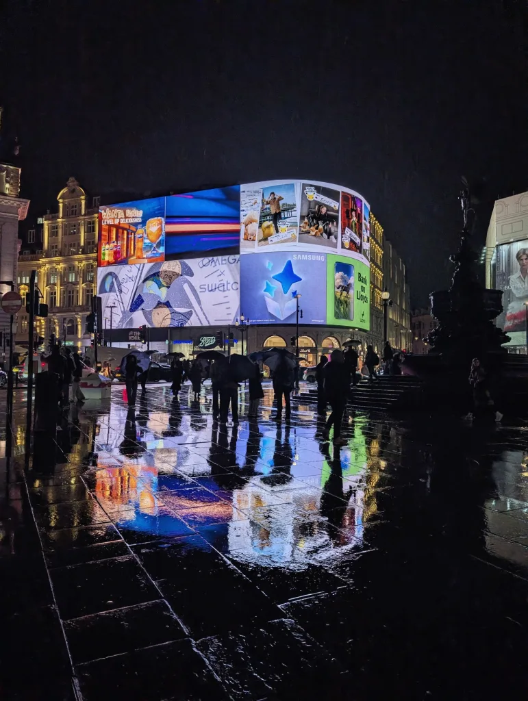 Pedestrians with umbrellas walk through Piccadilly Circus at night, with neon advertisements reflecting on the rain-soaked pavement.
