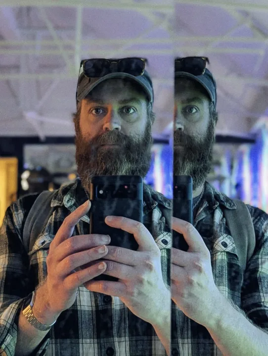 Jay Springett taking a mirrored selfie, bearded with sunglasses on cap, holding a phone, colourful bokeh lights behind