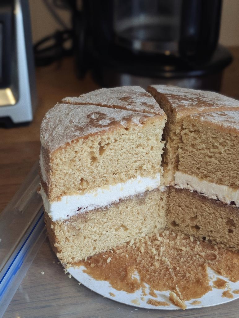 A Victoria sponge cake with cream and jam filling, dusted with icing sugar and sliced into wedges on a cardboard base.