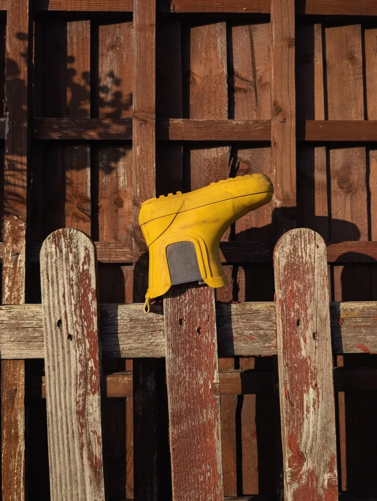 A bright yellow rubber boot rests upside down on a weathered wooden fence post in the sunlight against a brown trellis.