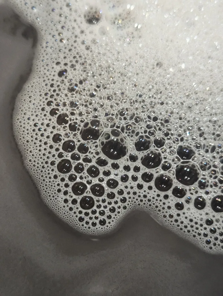 Dense white soap foam transitioning into larger translucent bubbles with iridescent glints over a dark grey surface.