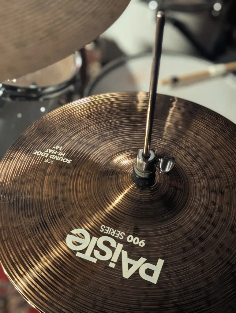 A Paiste 900 Series 14-inch Sound Edge hi-hat cymbal mounted on its stand, with other drums blurred in the warm background.