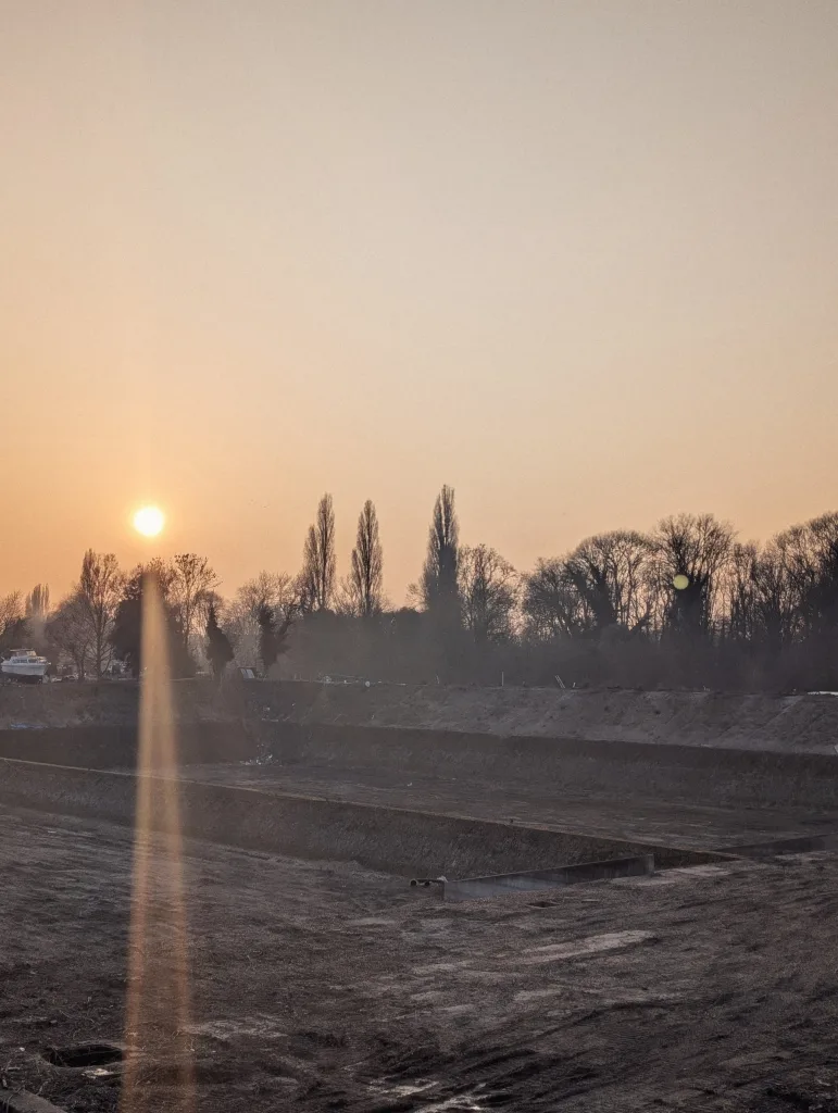 Low sun casting a lens flare over an empty concrete basin at a construction site, with silhouetted trees against a hazy orange sky.