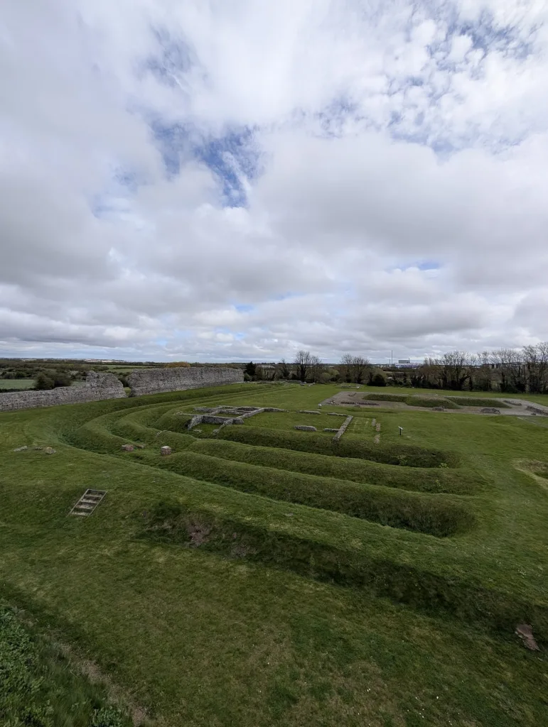 Low stone ruins and terraced grassy earthworks of Richborough Castle under a wide, cloudy sky.